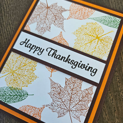 A close-up of a handmade Thanksgiving greeting card featuring a white background with stamped multicolor autumn leaves in yellow, orange, brown, and green. A dark brown banner across the center reads 'Happy Thanksgiving' in black script.