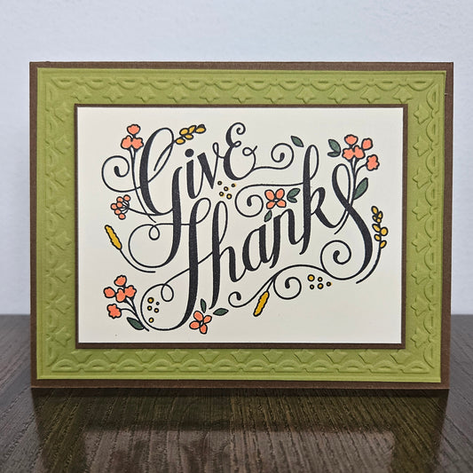 A framed handmade greeting card with the words 'Give Thanks' in black cursive script, decorated with small orange flowers and green leaves. The card has an off-white center, a textured olive-green inner border, and a dark brown outer frame, displayed on a dark wooden surface against a light gray wall.