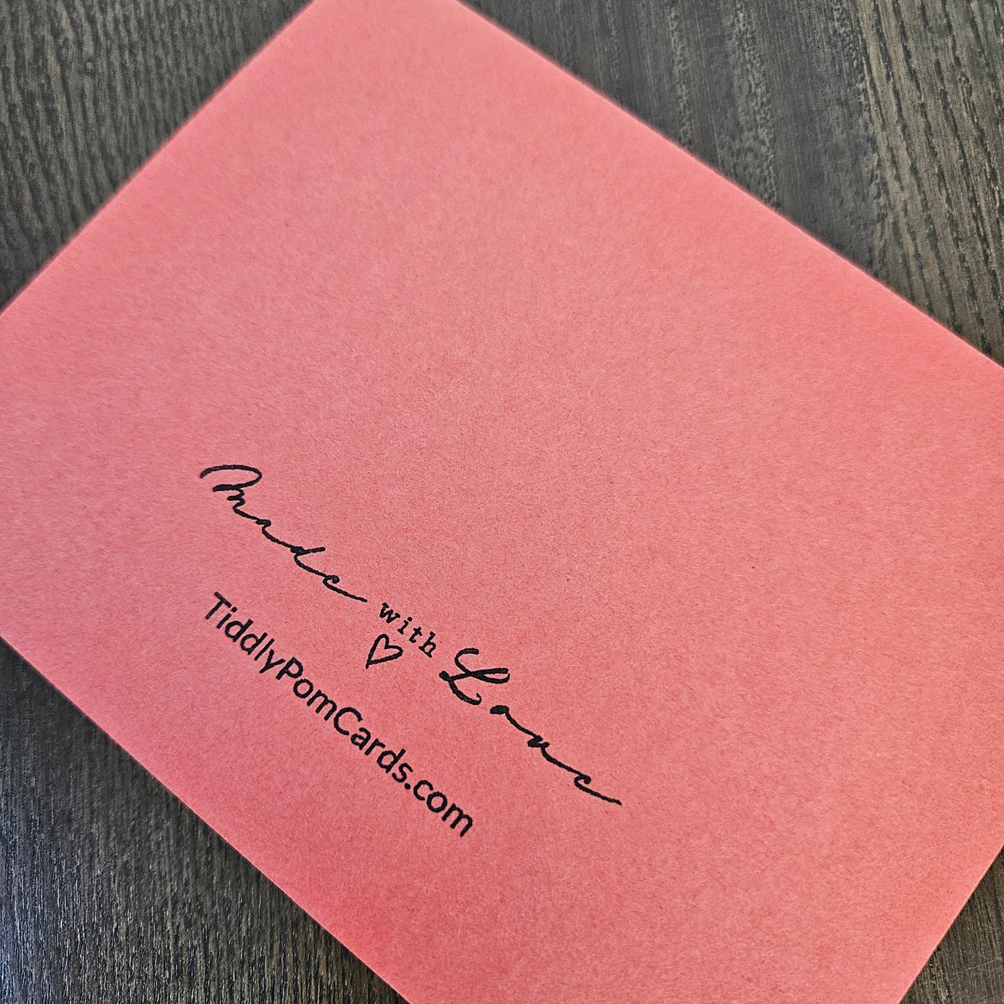 A coral pink textured greeting card with black printed cursive text reading 'Love with TiddlyPomCards.com' on a dark wood background.