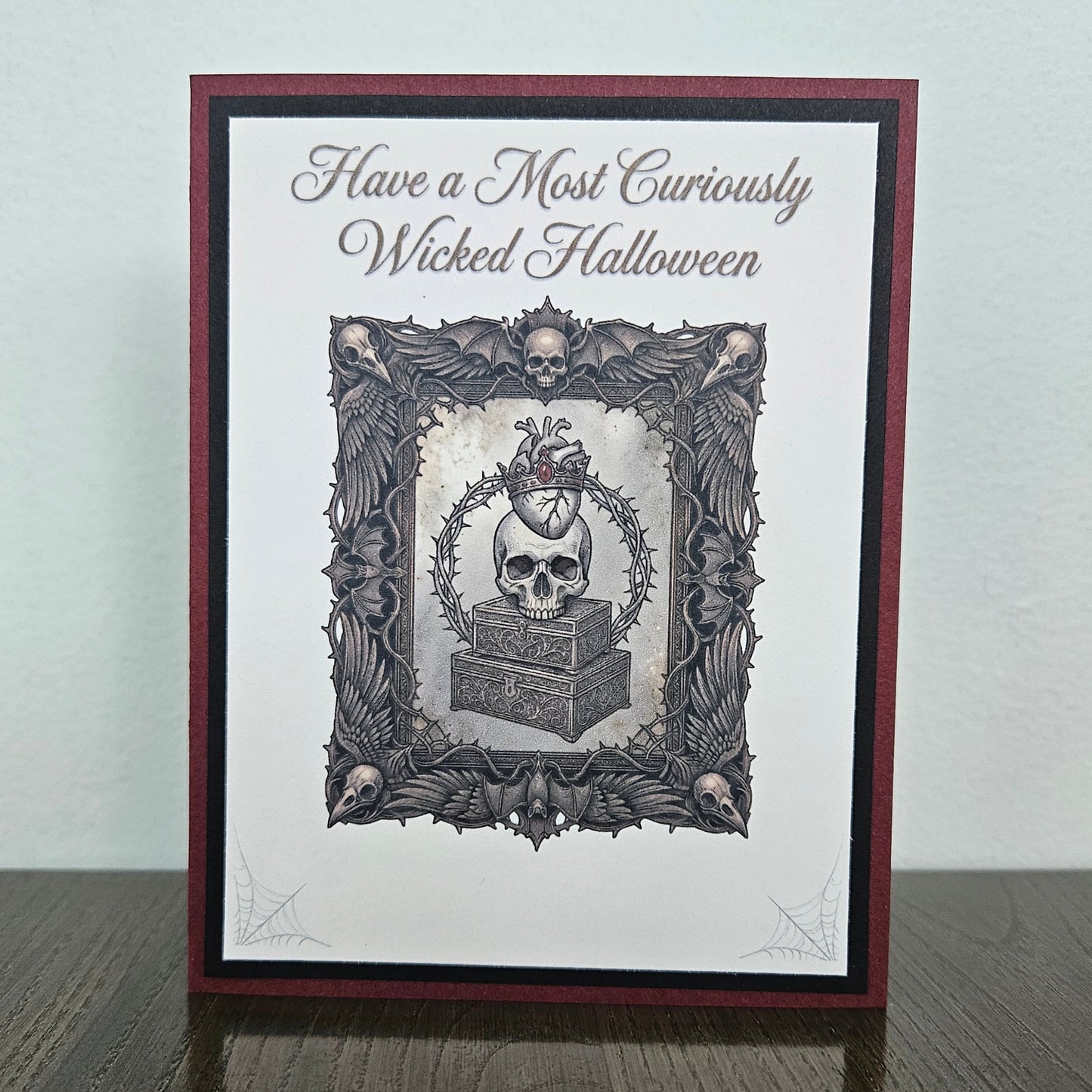 A handmade gothic-themed greeting card standing upright on a dark wooden surface against a light gray wall. The card features a dark red and black border, a white center, and a detailed black and gray illustration of a skull on a coffin inside an ornate frame, with the text 'Have a Most Curiously Wicked Halloween' at the top.