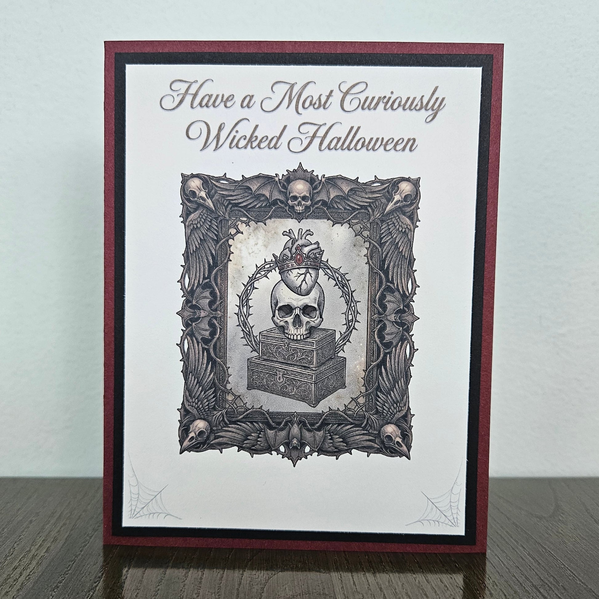 A handmade gothic-themed greeting card standing upright on a dark wooden surface against a light gray wall. The card features a dark red and black border, a white center, and a detailed black and gray illustration of a skull on a coffin inside an ornate frame, with the text 'Have a Most Curiously Wicked Halloween' at the top.