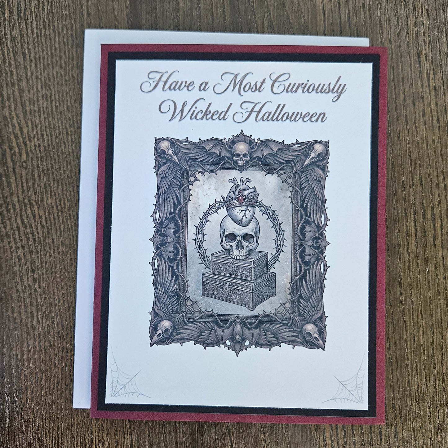 A square greeting card with a gothic Halloween design, featuring the text 'Have a Most Curiously Wicked Halloween' above an illustration of a skull in a decorative frame, placed on a wooden surface.
