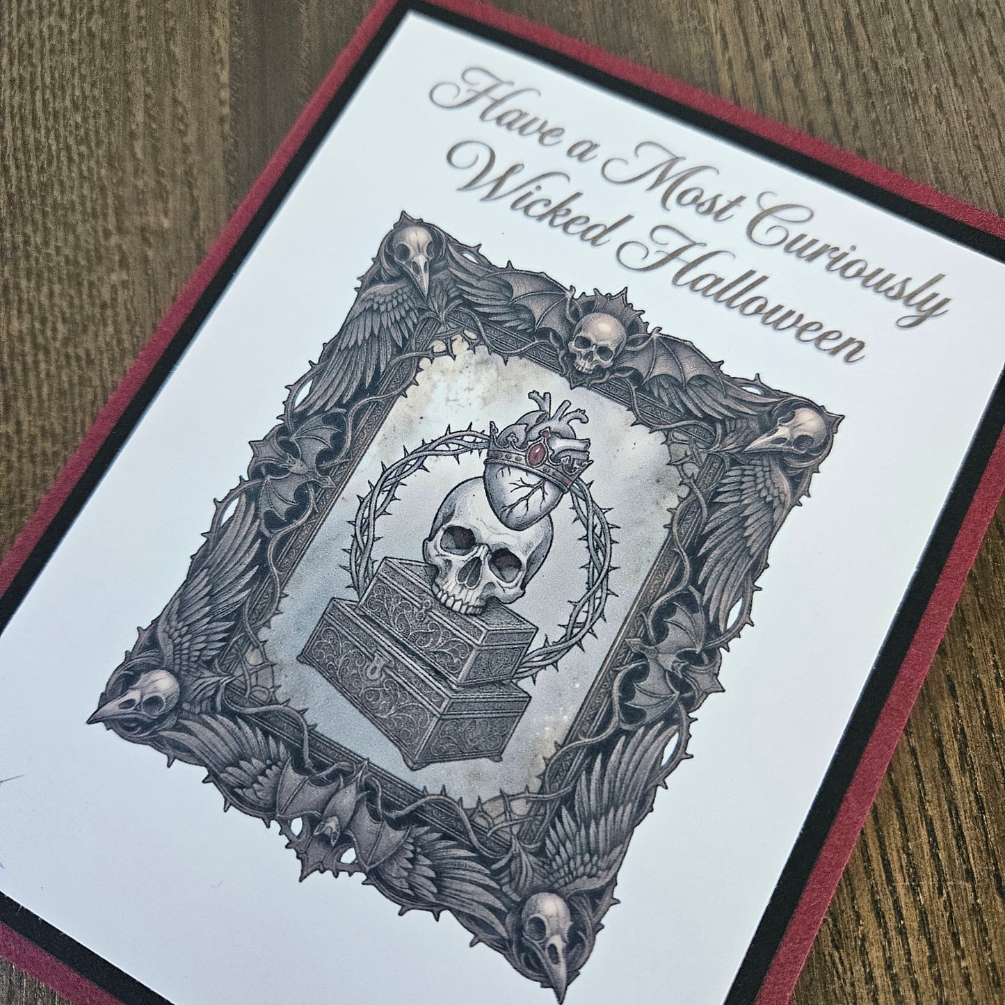 A close-up of a gothic-themed greeting card featuring a skull, heart, and ornate bat-and-skull border, with the text 'Have a Most Curiously Wicked Halloween' on a white card with a dark red and black border, placed on a wooden surface.