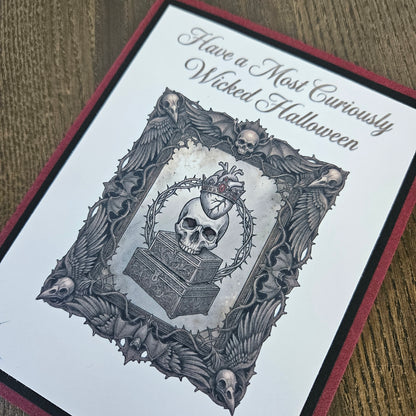A close-up of a gothic-themed greeting card featuring a skull, heart, and ornate bat-and-skull border, with the text 'Have a Most Curiously Wicked Halloween' on a white card with a dark red and black border, placed on a wooden surface.
