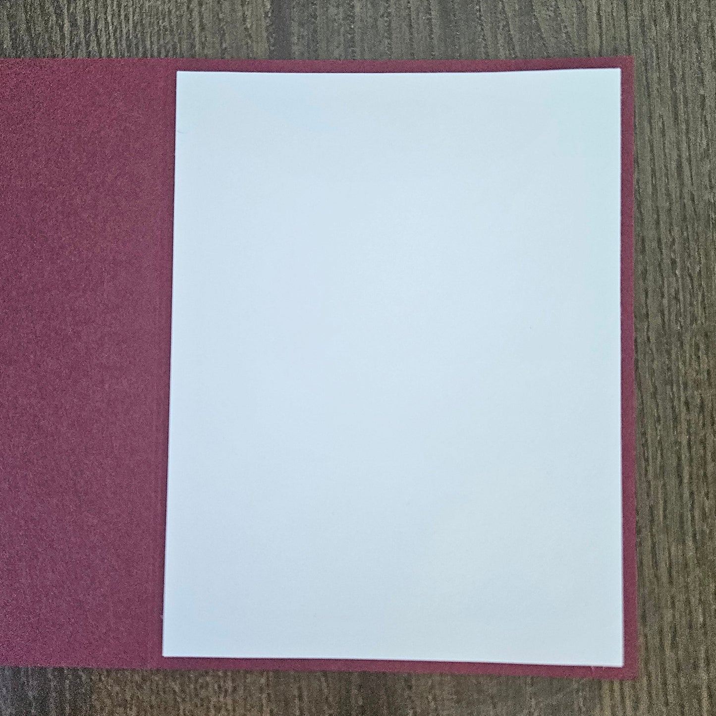 A blank white sheet of paper centered on a textured burgundy or deep red rectangular card or folder, placed on a brown wood-grain surface.