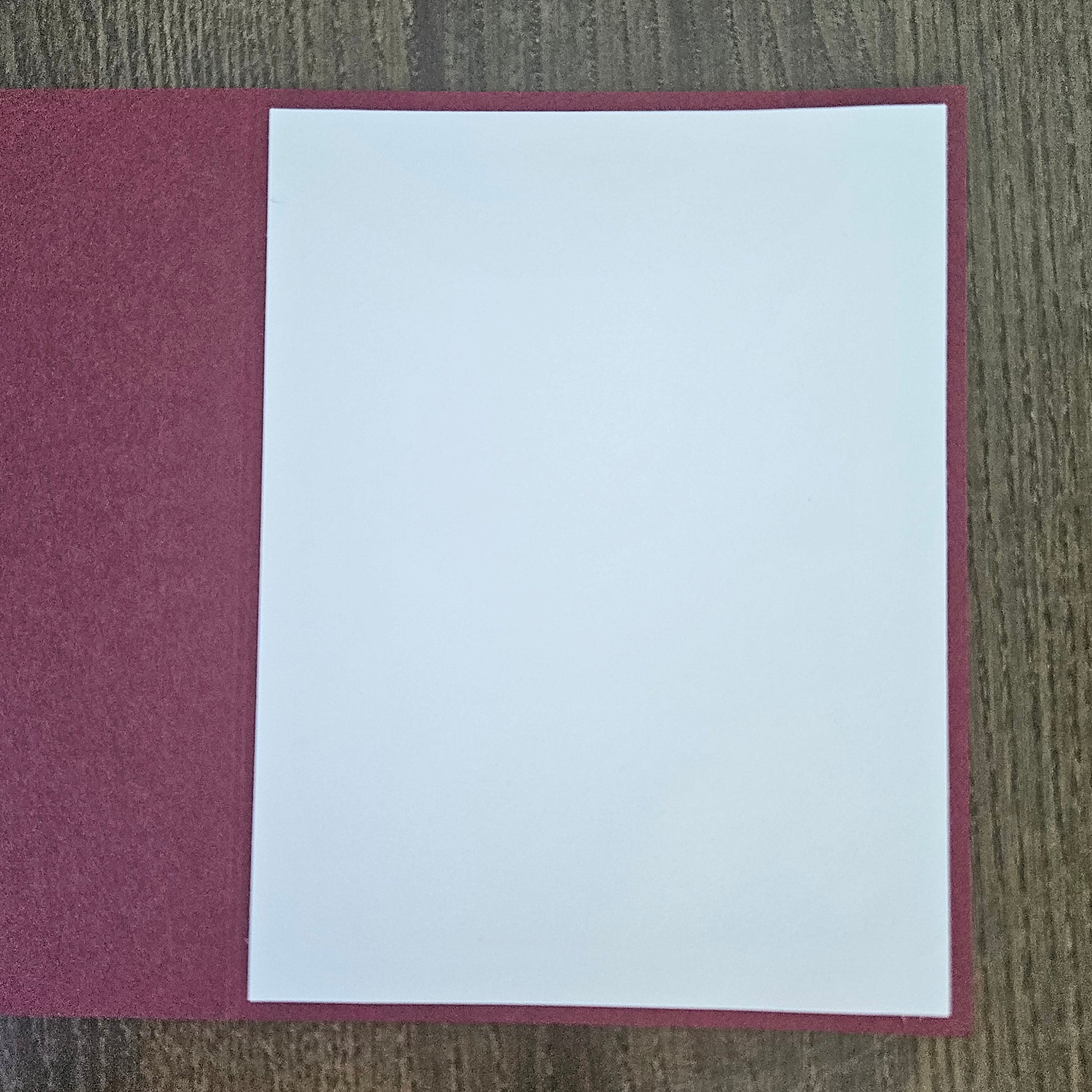 A blank white sheet of paper centered on a textured burgundy or deep red rectangular card or folder, placed on a brown wood-grain surface.