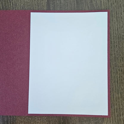 A blank white sheet of paper centered on a textured burgundy or deep red rectangular card or folder, placed on a brown wood-grain surface.