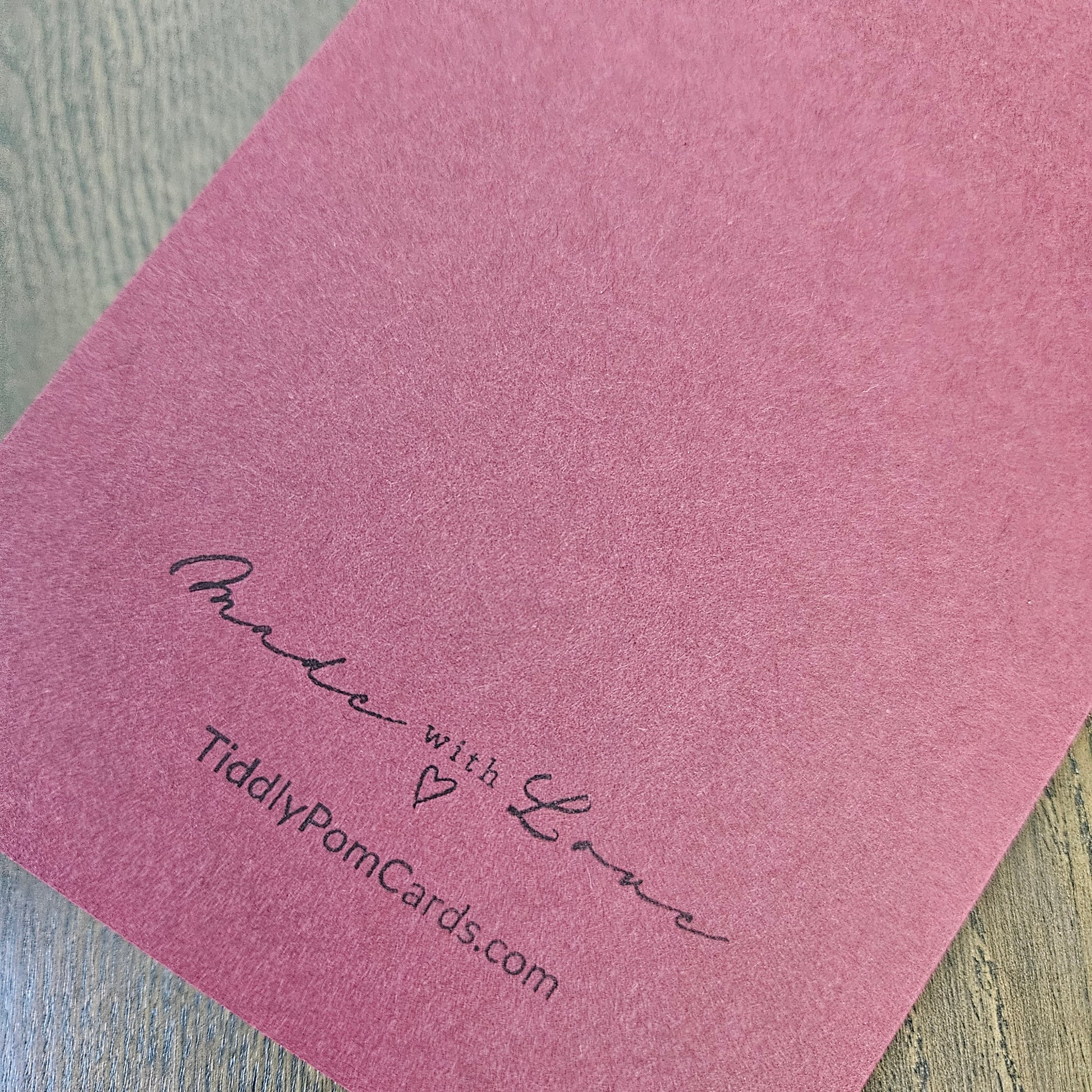 A close-up of a dusty rose pink textured card featuring the printed text 'Made with Love' and 'TiddlyPomCards.com' in black script, resting on a light gray wood-grain surface.