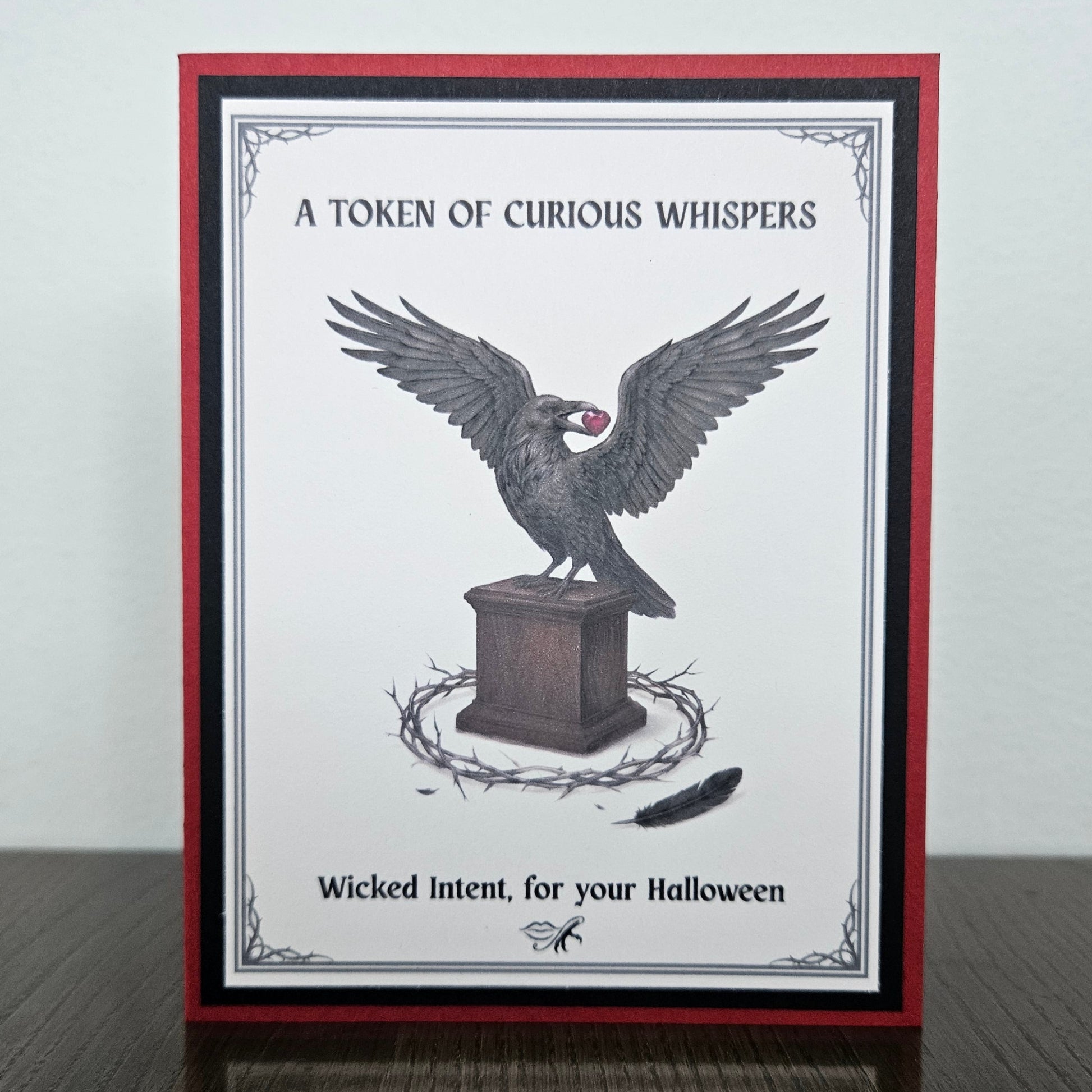 Front view of a handmade A2 gothic Halloween card featuring a raven holding a heart on a pedestal surrounded by thorns with crimson red borders. 