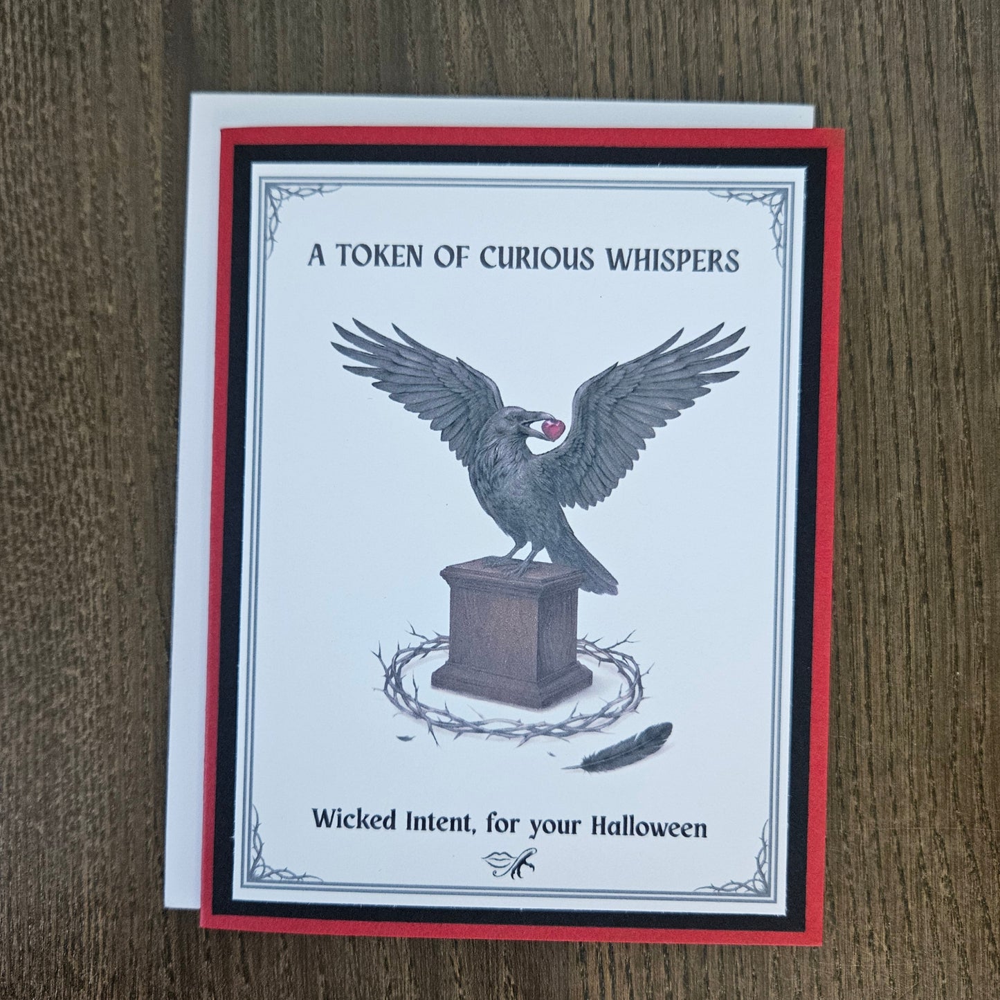 A square greeting card with a white center, red and black border, and a raven perched on a pedestal with the text 'A TOKEN OF CURIOUS WHISPERS' and 'Wicked Intent, for your Halloween,' photographed on a brown wood surface.
