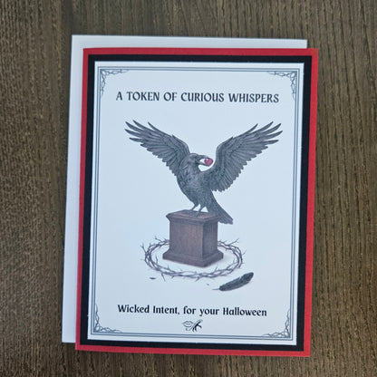 A square greeting card with a white center, red and black border, and a raven perched on a pedestal with the text 'A TOKEN OF CURIOUS WHISPERS' and 'Wicked Intent, for your Halloween,' photographed on a brown wood surface.