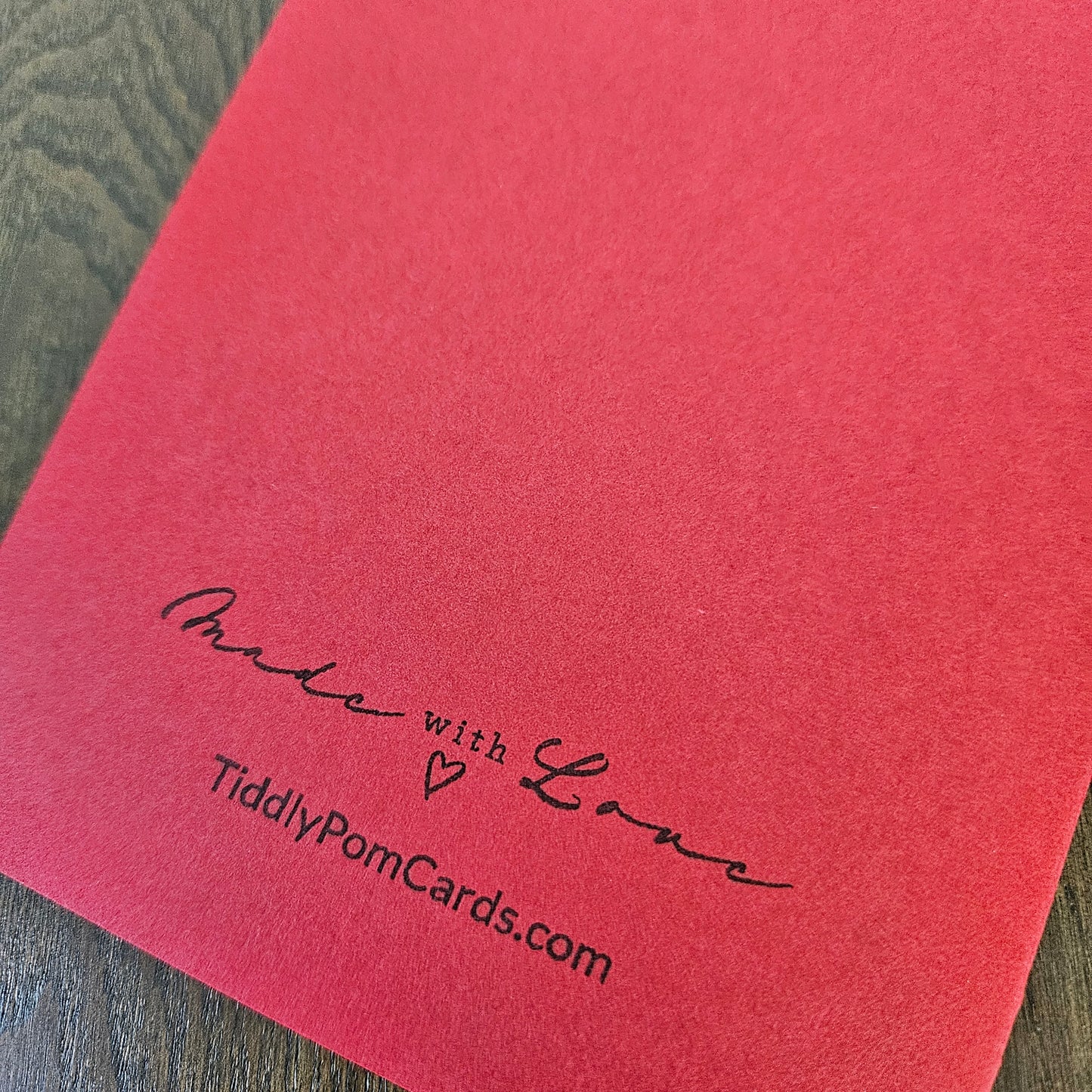 A close-up of a red textured card or envelope corner with black printed text reading 'Made with Love' and 'TiddlyPomCards.com' on a dark wood surface.