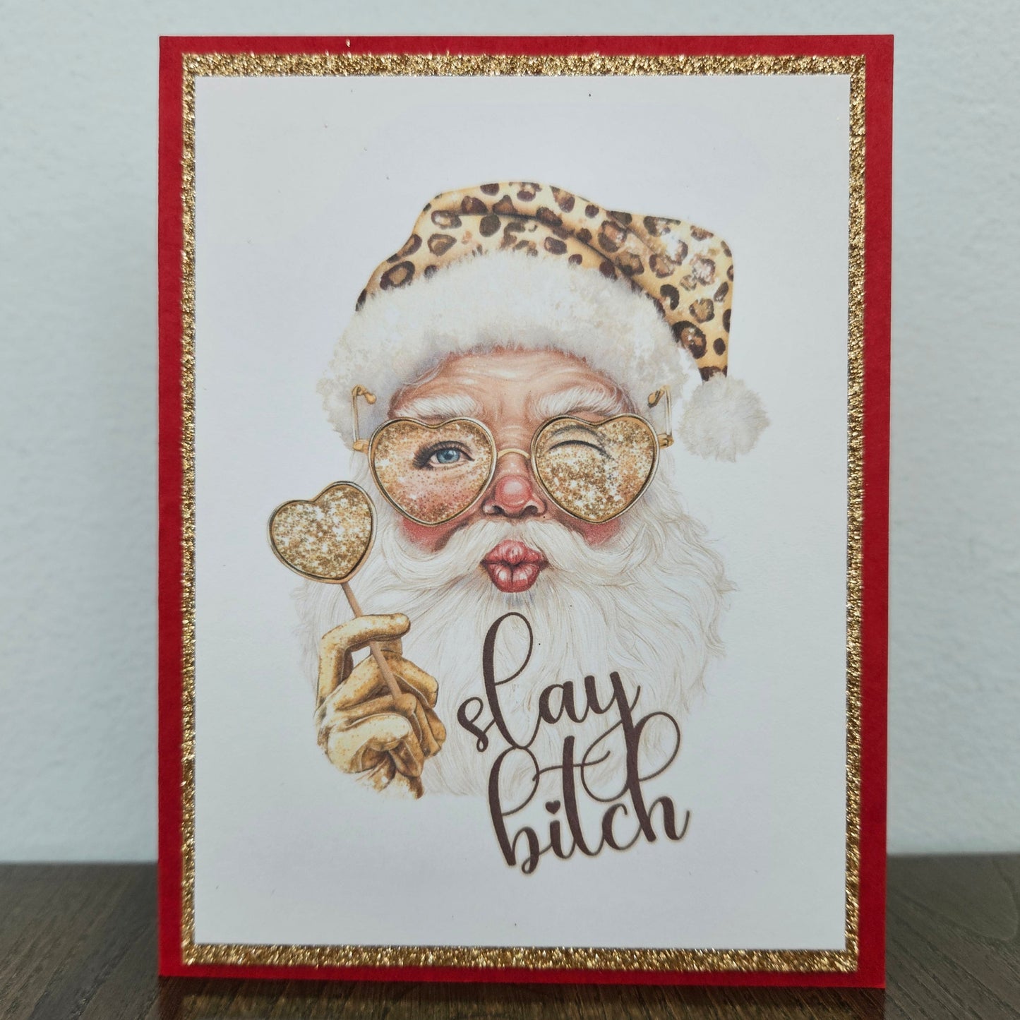 A handmade greeting card featuring a whimsical illustration of Santa Claus with a leopard-print hat, glittery round glasses, and a heart-shaped lollipop, with the text 'slay bitch' written below. The card has a red border with a glittery gold inner trim and is displayed standing on a dark wooden surface against a light gray wall.