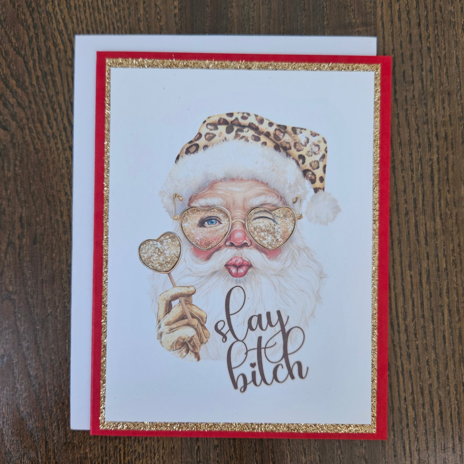 A square greeting card featuring a stylized illustration of Santa Claus with a leopard-print hat, heart-shaped glasses, and a white beard, holding a heart-shaped ornament. The card has a red border with glittery gold trim and the phrase 'slay bitch' in script.
