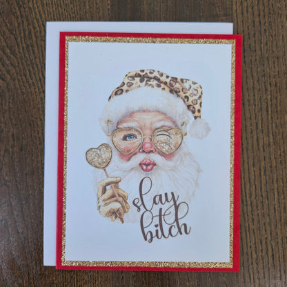 A square greeting card featuring a stylized illustration of Santa Claus with a leopard-print hat, heart-shaped glasses, and a white beard, holding a heart-shaped ornament. The card has a red border with glittery gold trim and the phrase 'slay bitch' in script.