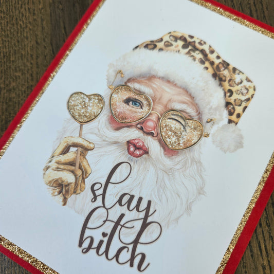 A close-up of a printed card or art piece featuring an illustrated Santa Claus with a leopard-print Santa hat, heart-shaped glittery glasses, a white beard, and the text 'stay bitch' in dark script. The design has a red border with gold glitter trim and is photographed on a wooden surface.