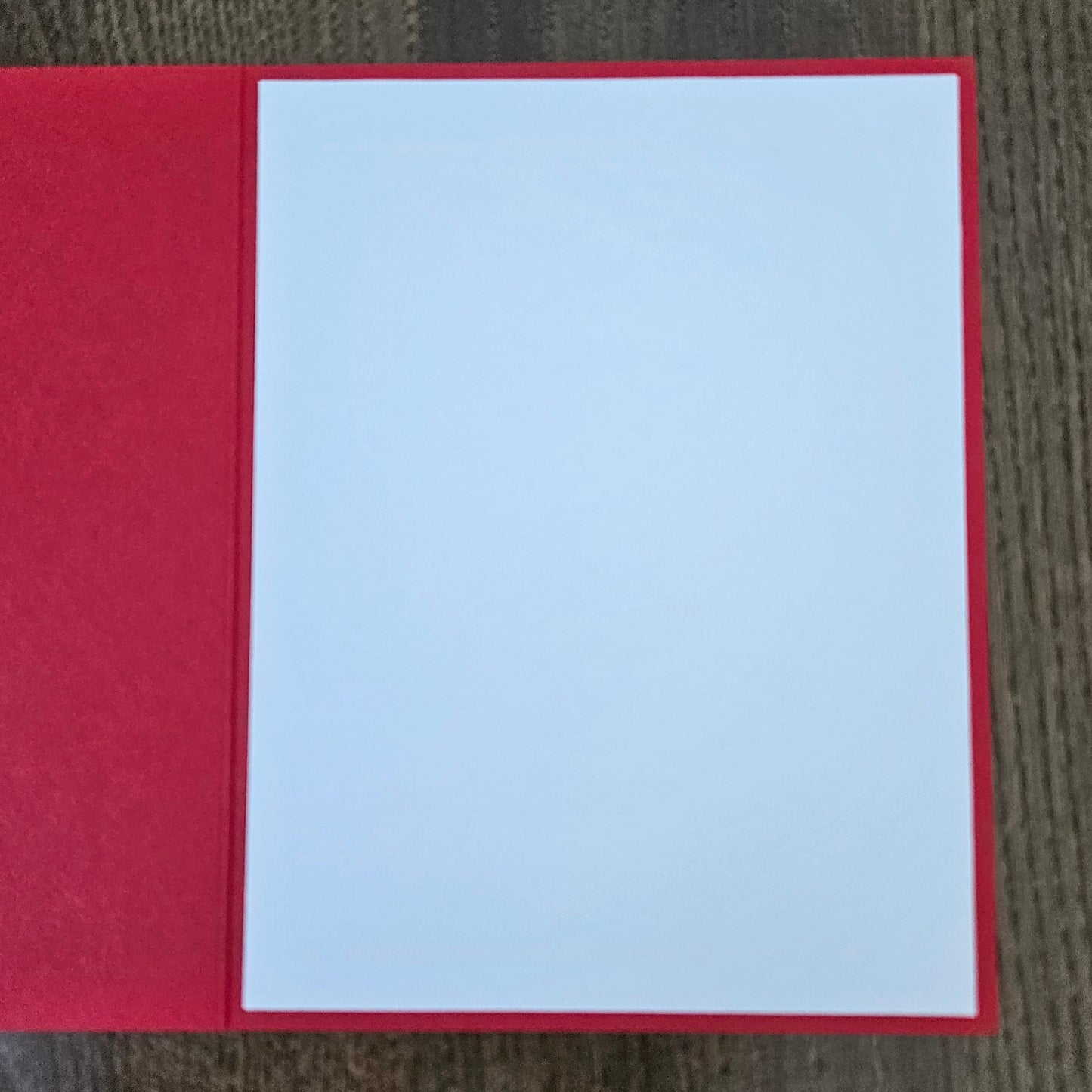 A red greeting card or notebook cover with a blank white center panel, photographed from above on a dark wood surface.