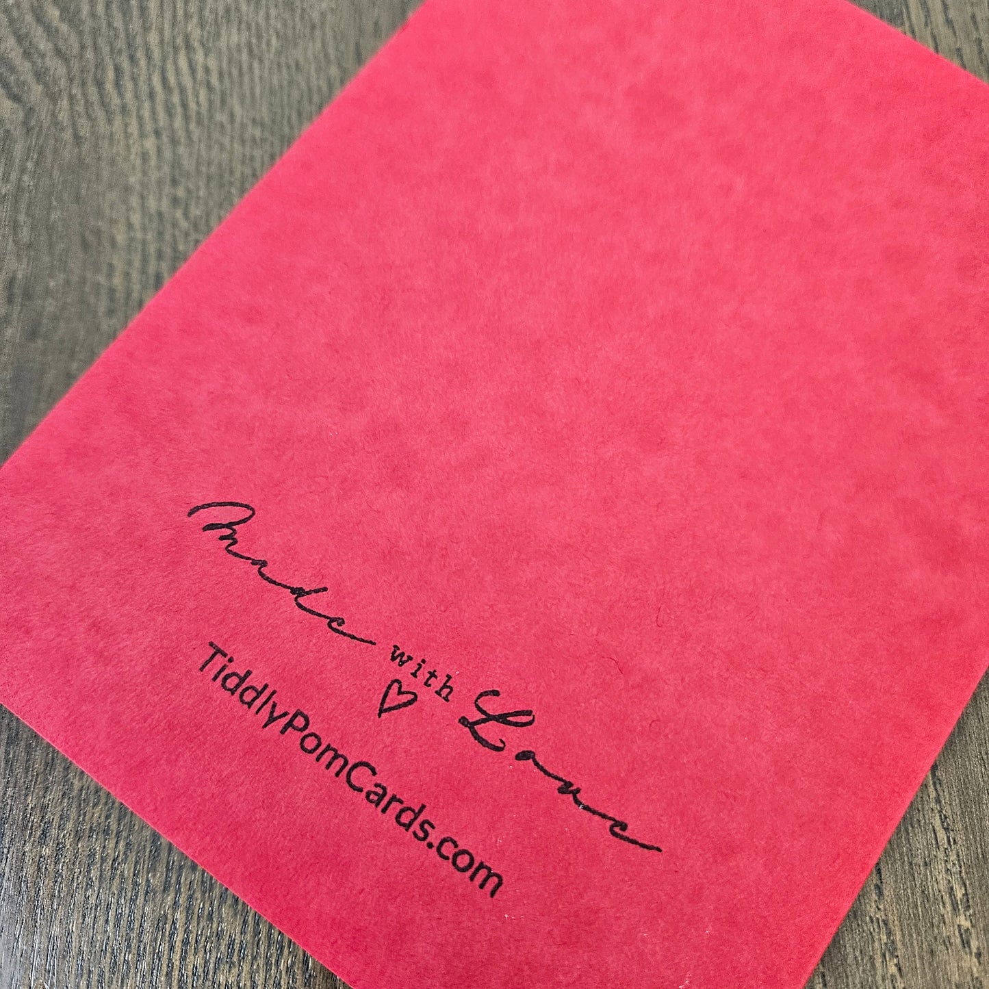 A close-up of a bright pink textured card or paper sheet placed on a gray wood-grain surface, with black printed text reading 'Make with Love' and 'TiddlyPomCards.com'.