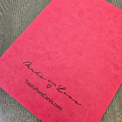 A close-up of a bright pink textured card or paper sheet placed on a gray wood-grain surface, with black printed text reading 'Make with Love' and 'TiddlyPomCards.com'.