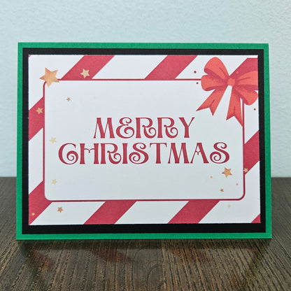 A handmade Christmas greeting card with 'MERRY CHRISTMAS' in red script, featuring red and white diagonal stripes, a red bow, and gold stars, set against a green and black border on a wooden surface.