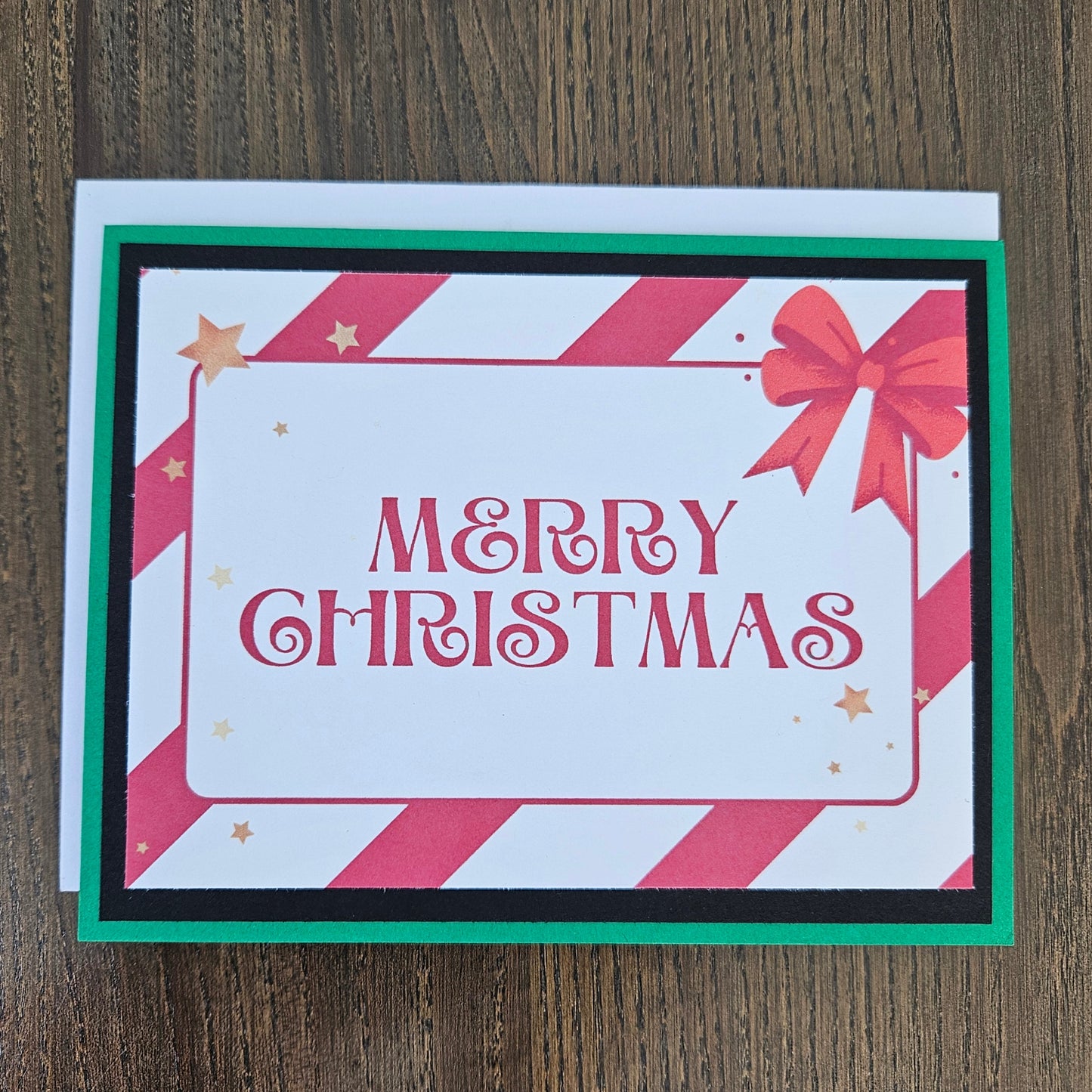 A square Christmas greeting card with 'MERRY CHRISTMAS' in red script, decorated with red and white diagonal stripes, a red bow, and small gold stars. The card is layered with black and green borders and sits on a white envelope over a dark wood background.