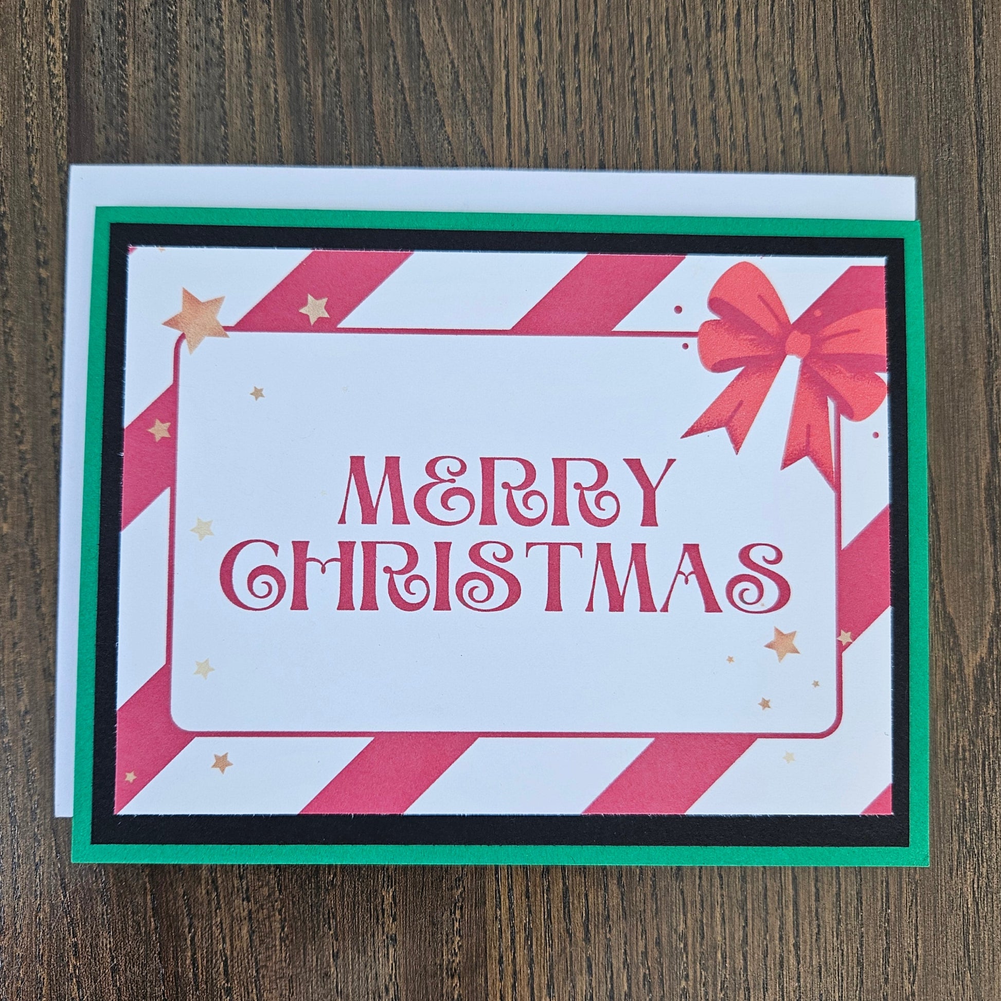 A square Christmas greeting card with 'MERRY CHRISTMAS' in red script, decorated with red and white diagonal stripes, a red bow, and small gold stars. The card is layered with black and green borders and sits on a white envelope over a dark wood background.