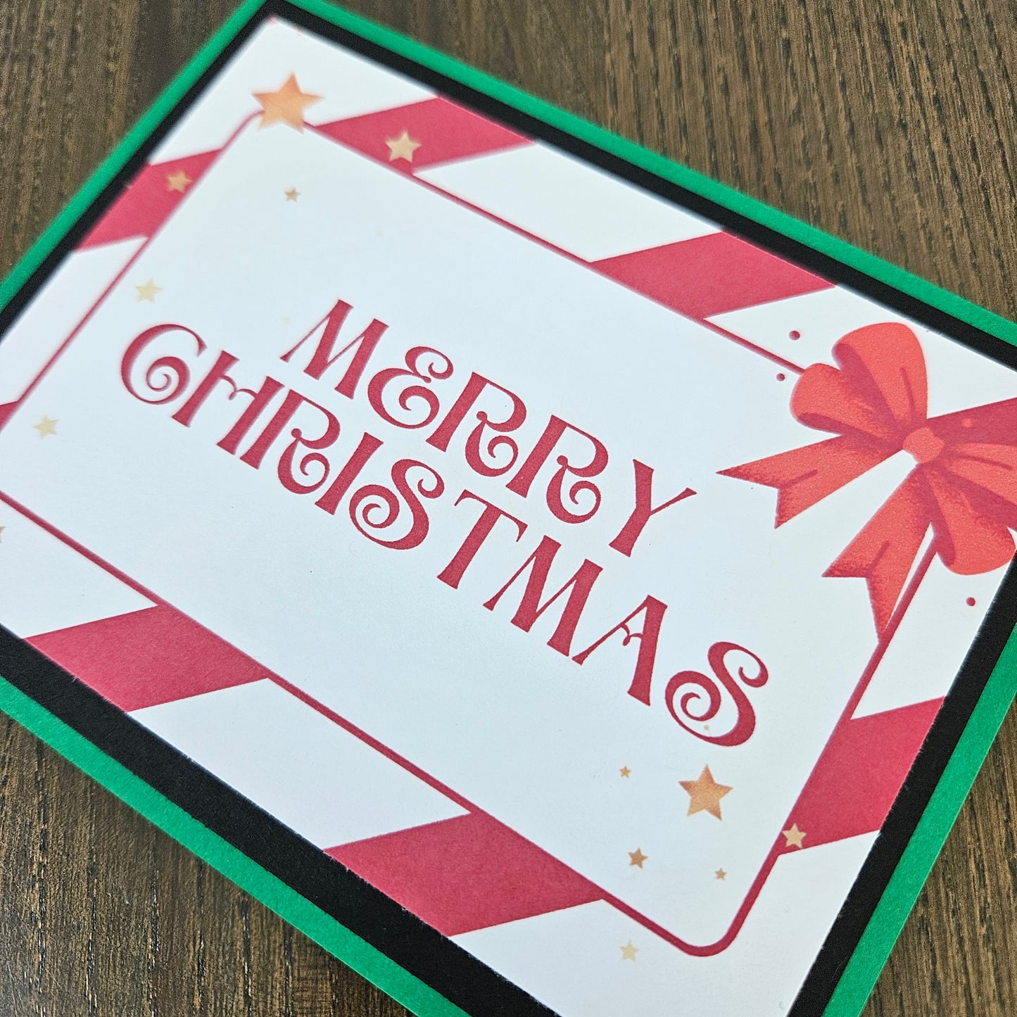 A square Christmas greeting card with 'MERRY CHRISTMAS' in red script, a red bow illustration, and red-and-white diagonal stripes around a white center panel, photographed on a wooden surface.