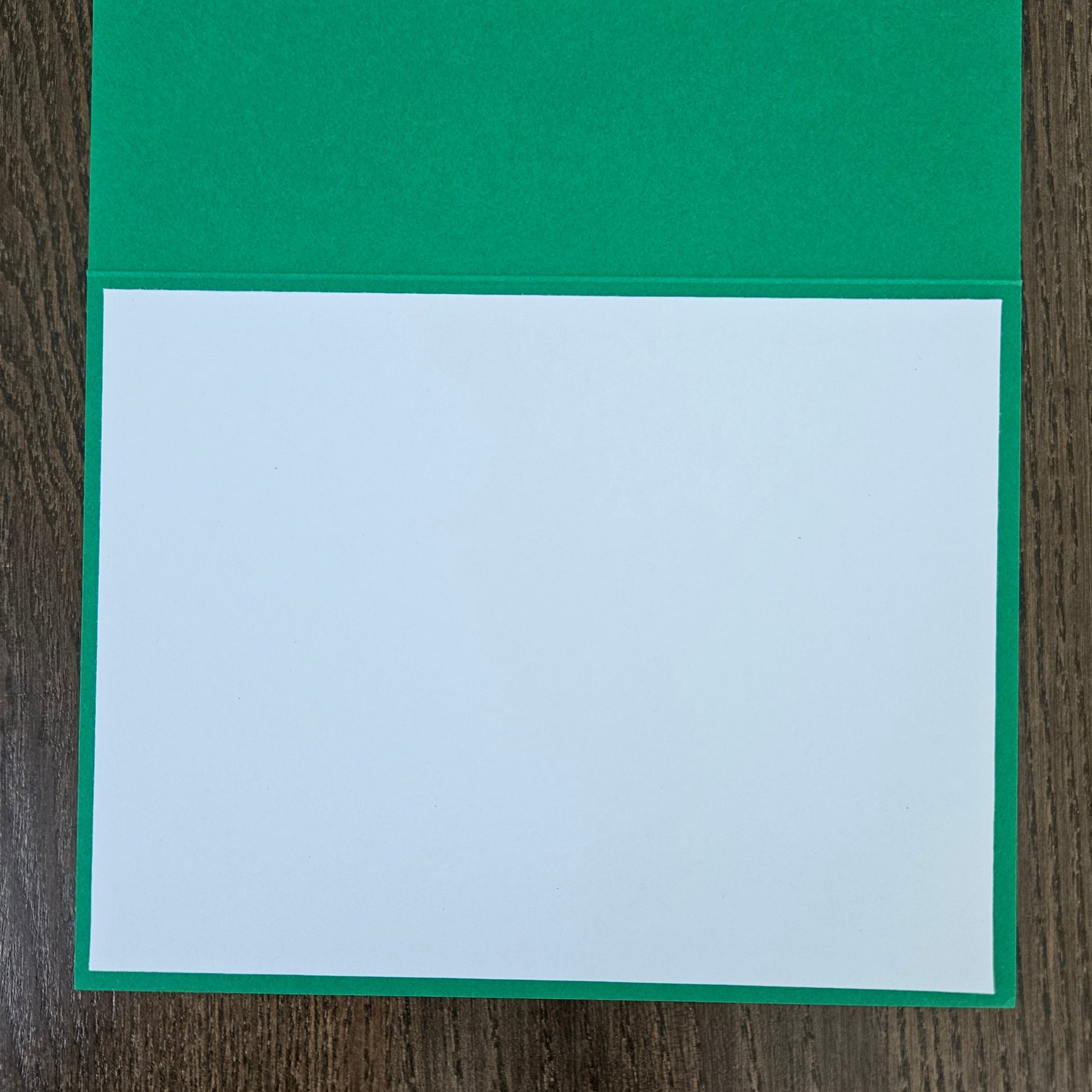 A green greeting card with a blank white center panel, displayed flat on a dark wood background.