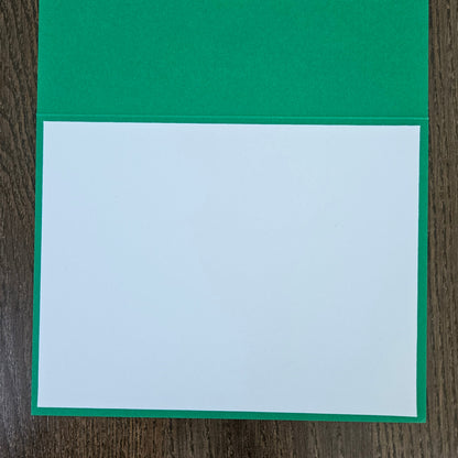 A green greeting card with a blank white center panel, displayed flat on a dark wood background.