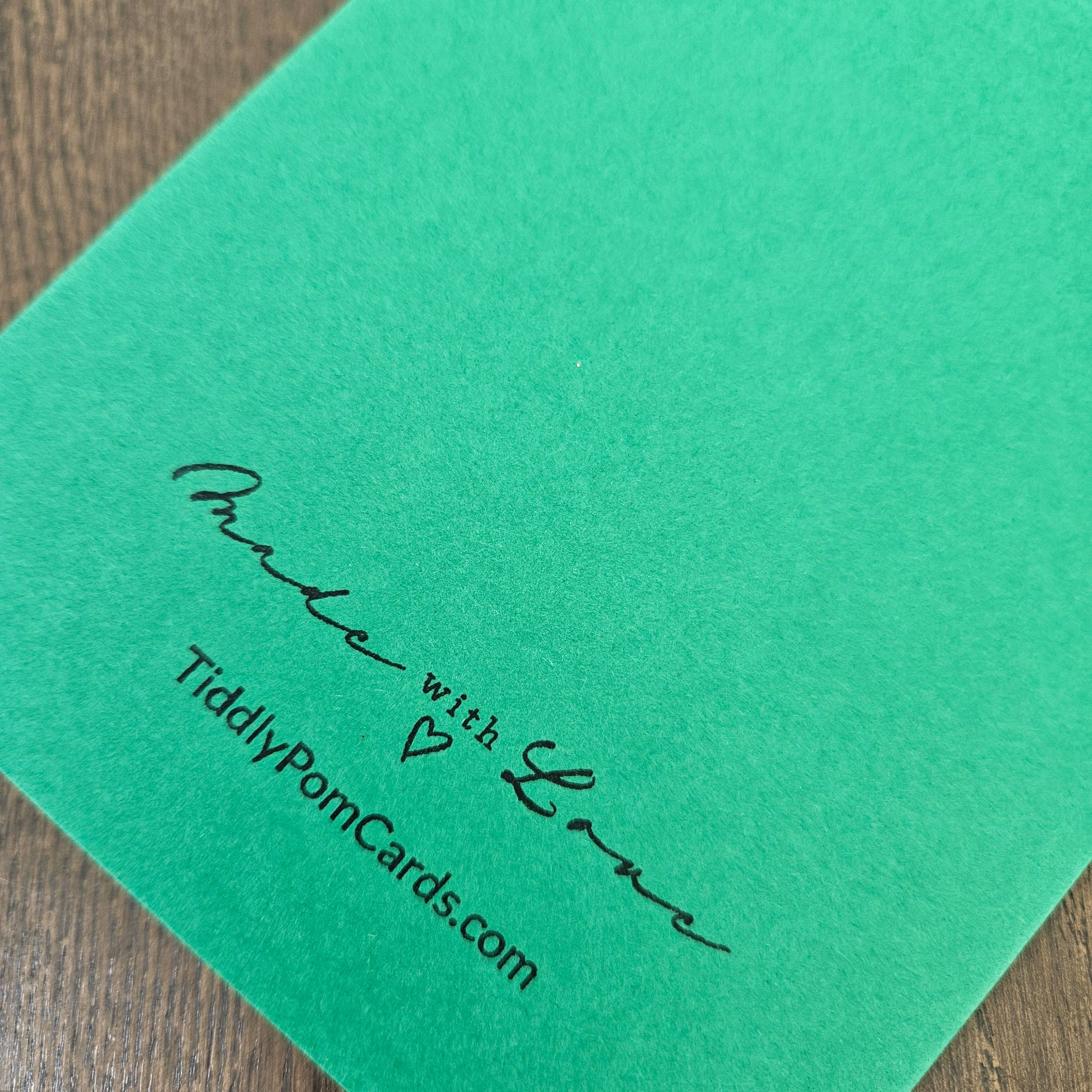A close-up of a bright green card featuring black printed text that reads 'Proud with Love' and 'TiddlyPomCards.com' on a wooden surface.