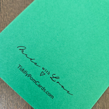 A close-up of a bright green card featuring black printed text that reads 'Proud with Love' and 'TiddlyPomCards.com' on a wooden surface.