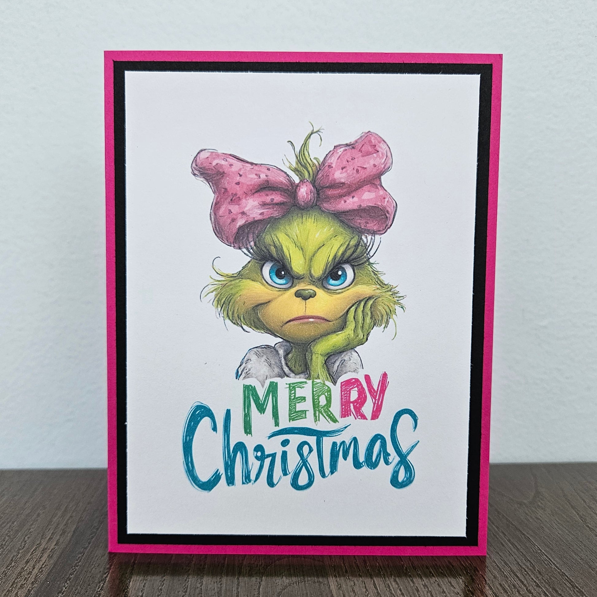 A handmade Christmas greeting card featuring an illustration of the Grinch with a pink bow and the text 'MERRY Christmas' in colorful lettering, set against a white background with a bright pink border.