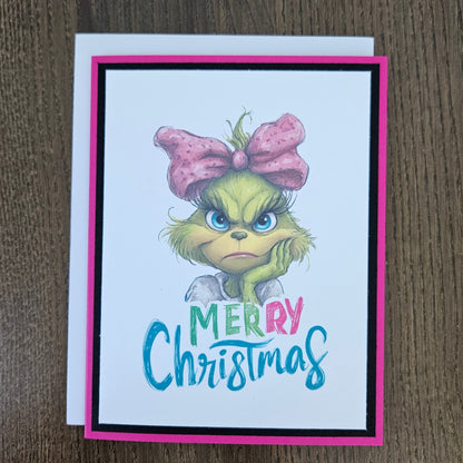 A handmade Christmas greeting card featuring a cartoon illustration of the Grinch with a pink bow and the text 'MERRY Christmas' in colorful lettering, displayed on a dark wood background.