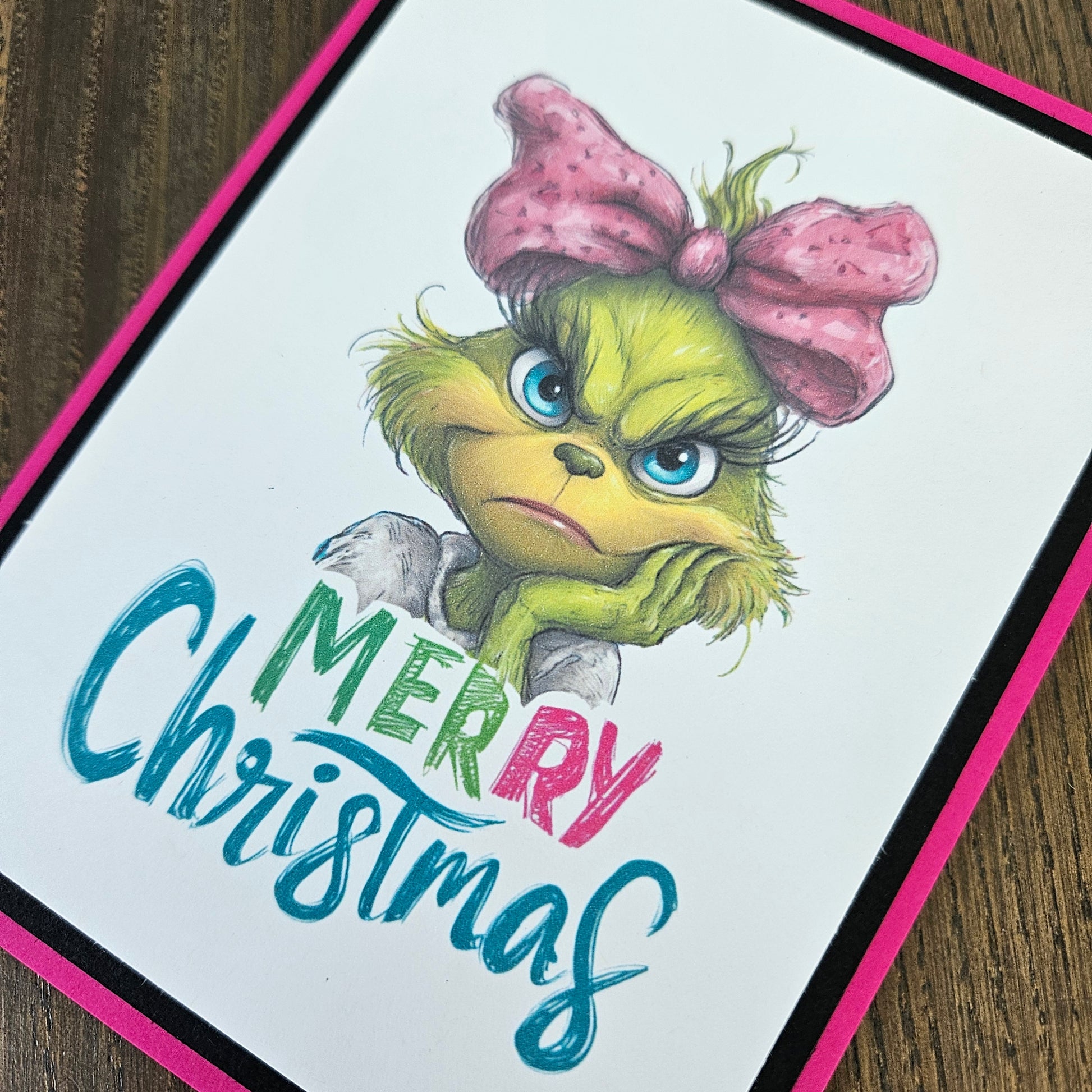 A square greeting card featuring an illustration of the Grinch with a pink bow and the text 'Merry Christmas' in colorful lettering, photographed on a wooden surface.