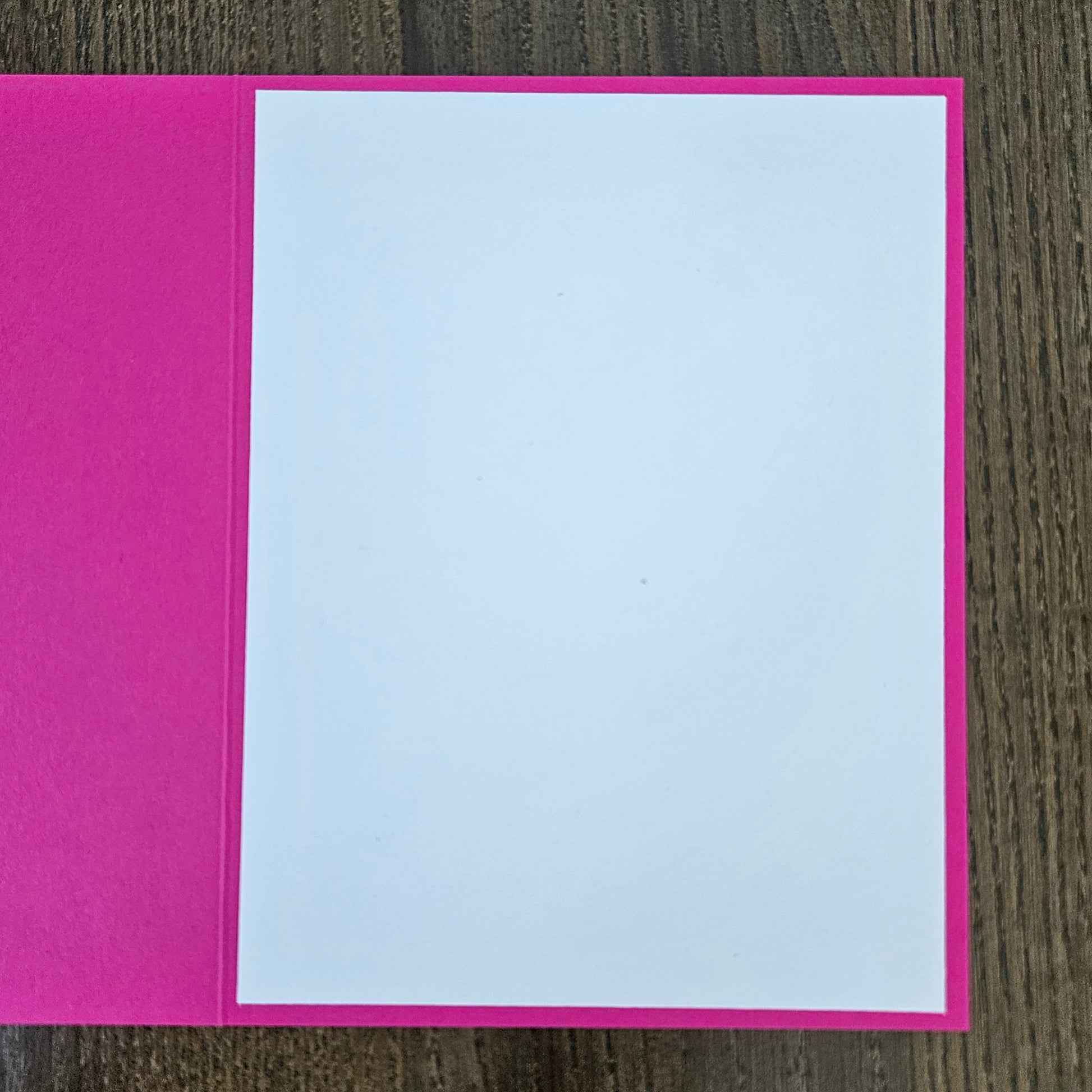 A blank greeting card with a bright pink cover and a white insert page, shown open on a dark wood background.