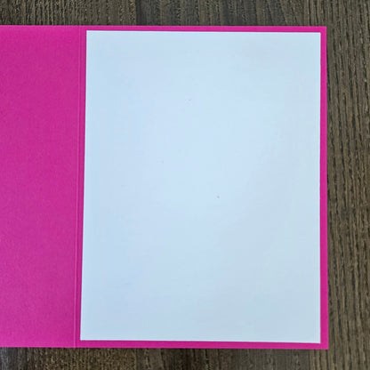 A blank greeting card with a bright pink cover and a white insert page, shown open on a dark wood background.