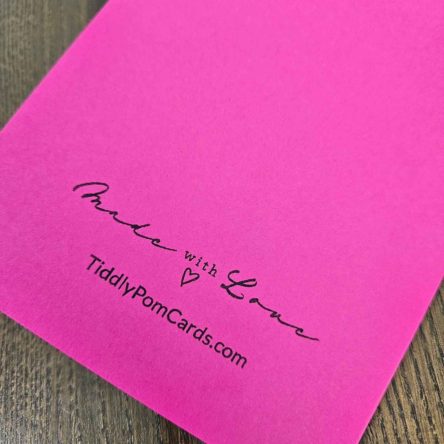 A bright pink square card or paper sheet with black printed text reading 'Made with Love' and 'TiddlyPomCards.com', photographed on a wood surface.