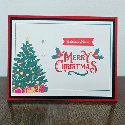 A handmade Christmas greeting card with a red and black layered border, featuring a decorated green Christmas tree, wrapped gifts, holly leaves, and the text 'Wishing You a Merry Christmas' on a white background, set on a wooden table against a light wall.