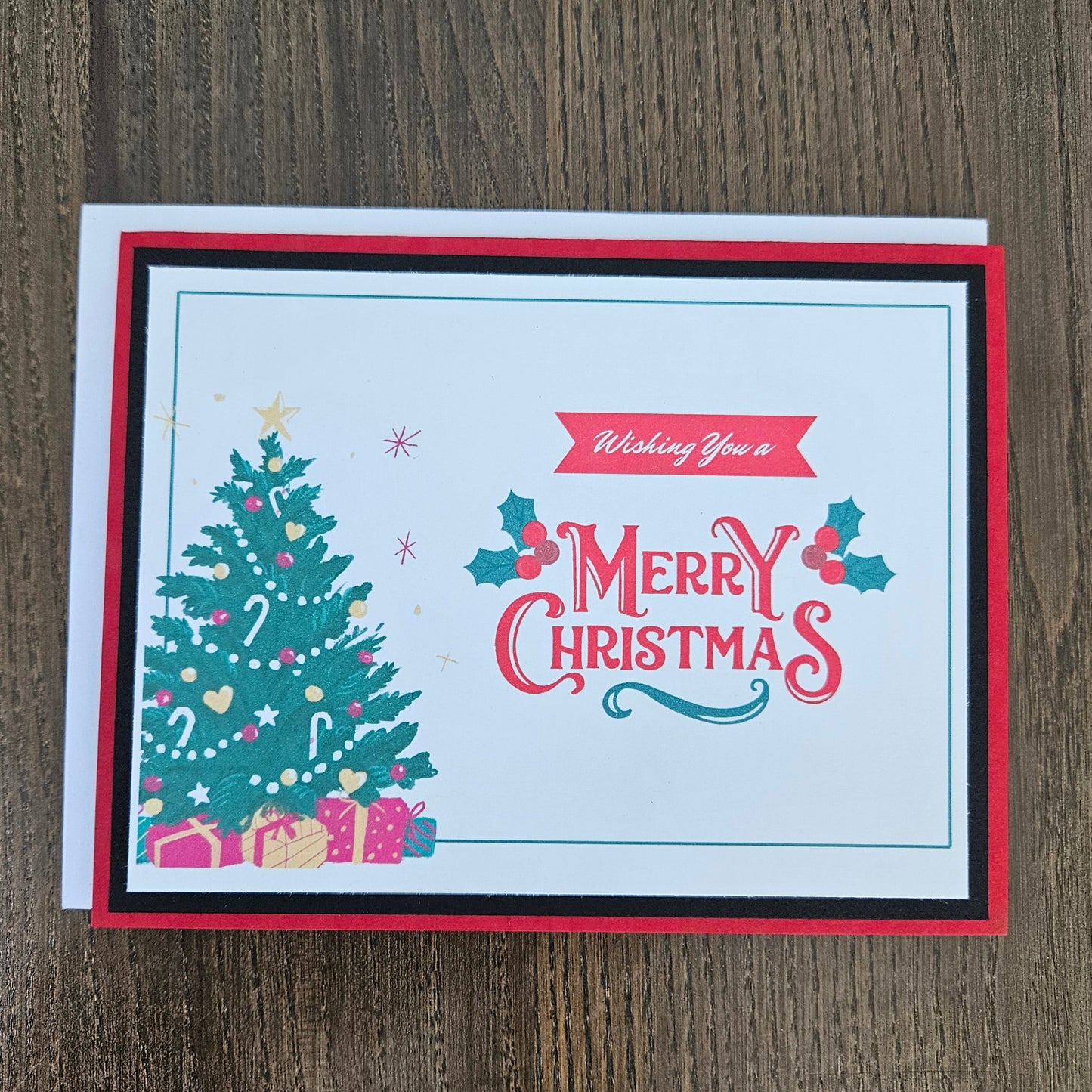 A square Christmas greeting card with a red, black, and white layered border, featuring a decorated green Christmas tree and the words 'Wishing You a Merry Christmas' in red and teal, placed on a dark wood background.