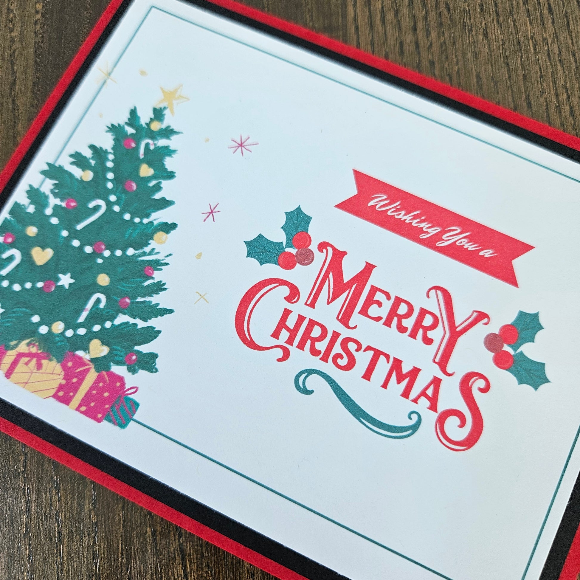 A square Christmas greeting card with a white center, red border, a green decorated tree, holly, candy canes, and the words 'Wishing You a Merry Christmas,' photographed on a wooden surface.