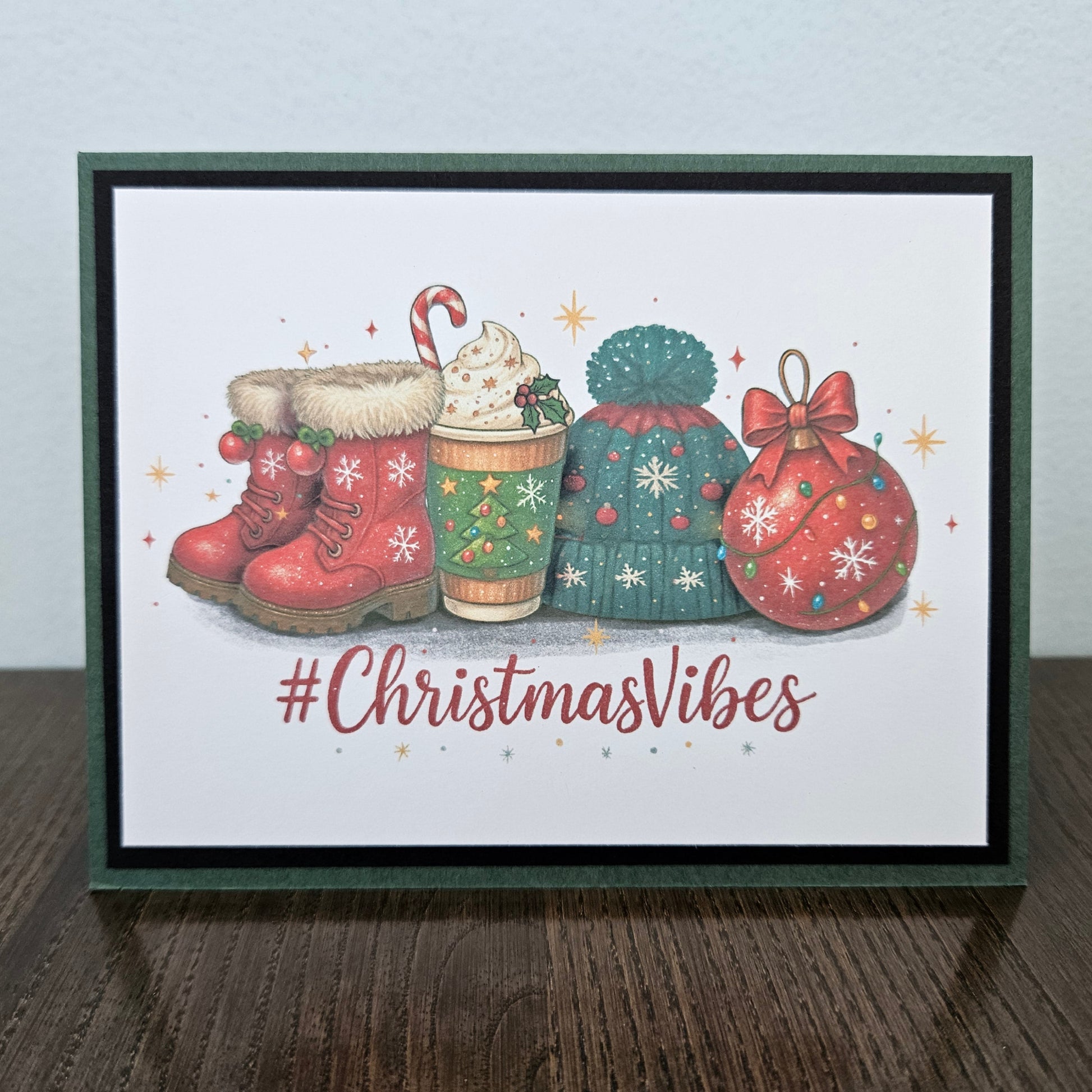 A framed Christmas greeting card on a dark wooden surface against a light gray wall. The card features an illustration of red winter boots, a hot cocoa cup, a green knitted hat, a red ornament, and the text '#ChristmasVibes' in red script.