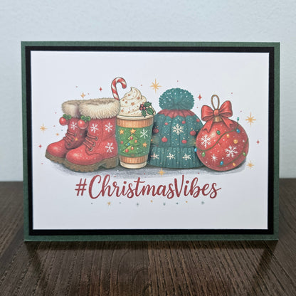 A framed Christmas greeting card on a dark wooden surface against a light gray wall. The card features an illustration of red winter boots, a hot cocoa cup, a green knitted hat, a red ornament, and the text '#ChristmasVibes' in red script.
