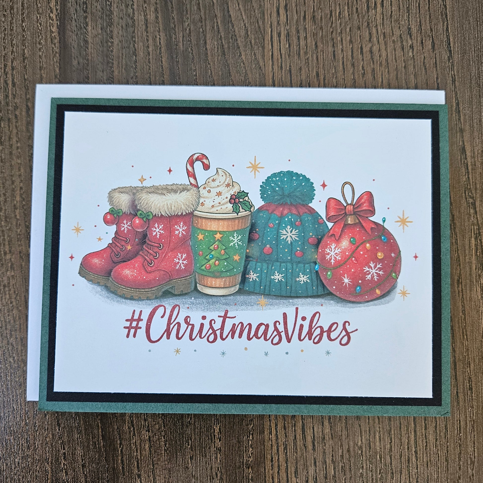 A square Christmas-themed greeting card featuring festive illustrations of red winter boots, a holiday drink with whipped cream and a candy cane, a teal knit hat, a red ornament, and the text '#ChristmasVibes' on a white card with a dark green and black border, placed on a wooden surface.