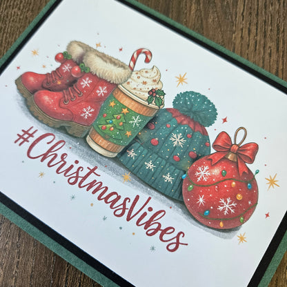 A square Christmas-themed card or print featuring illustrated red boots, a hot cocoa cup, a green knit hat, a red ornament, and the text '#ChristmasVibes' on a white background, photographed on a wooden surface.