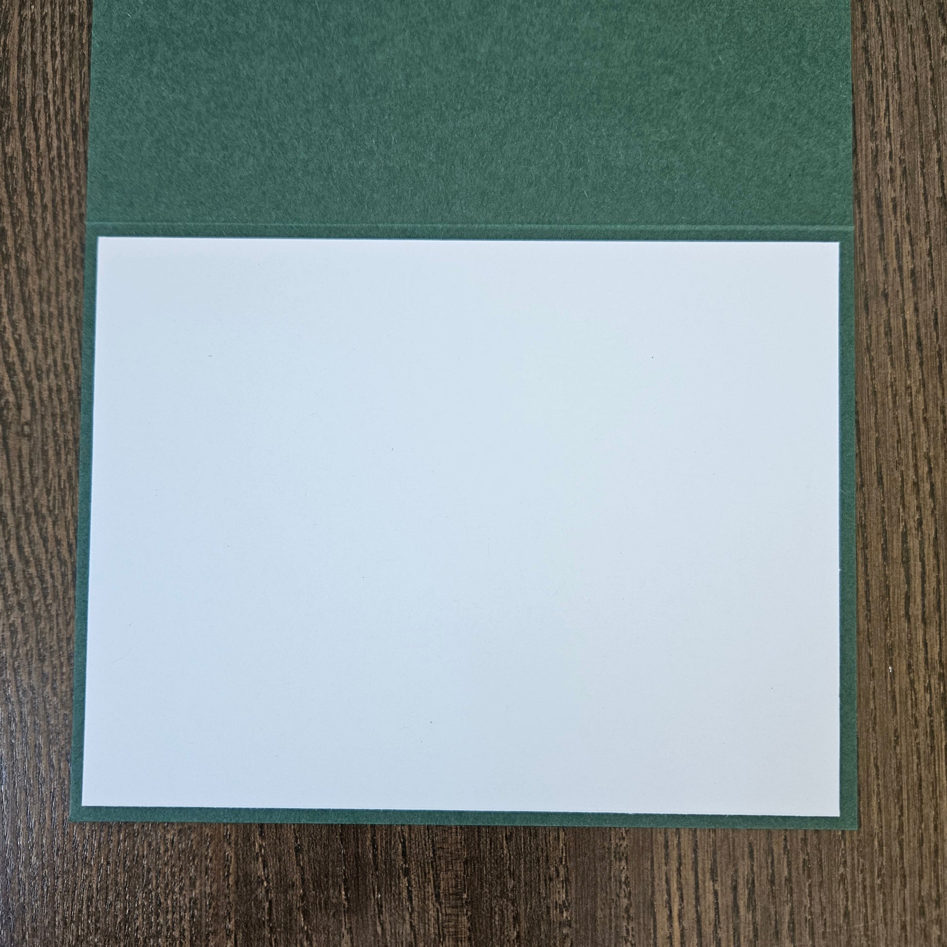 A square white card or paper insert placed on top of a dark green textured cover, photographed from above on a brown wood-grain surface.