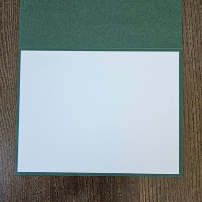 A square white card or paper insert placed on top of a dark green textured cover, photographed from above on a brown wood-grain surface.