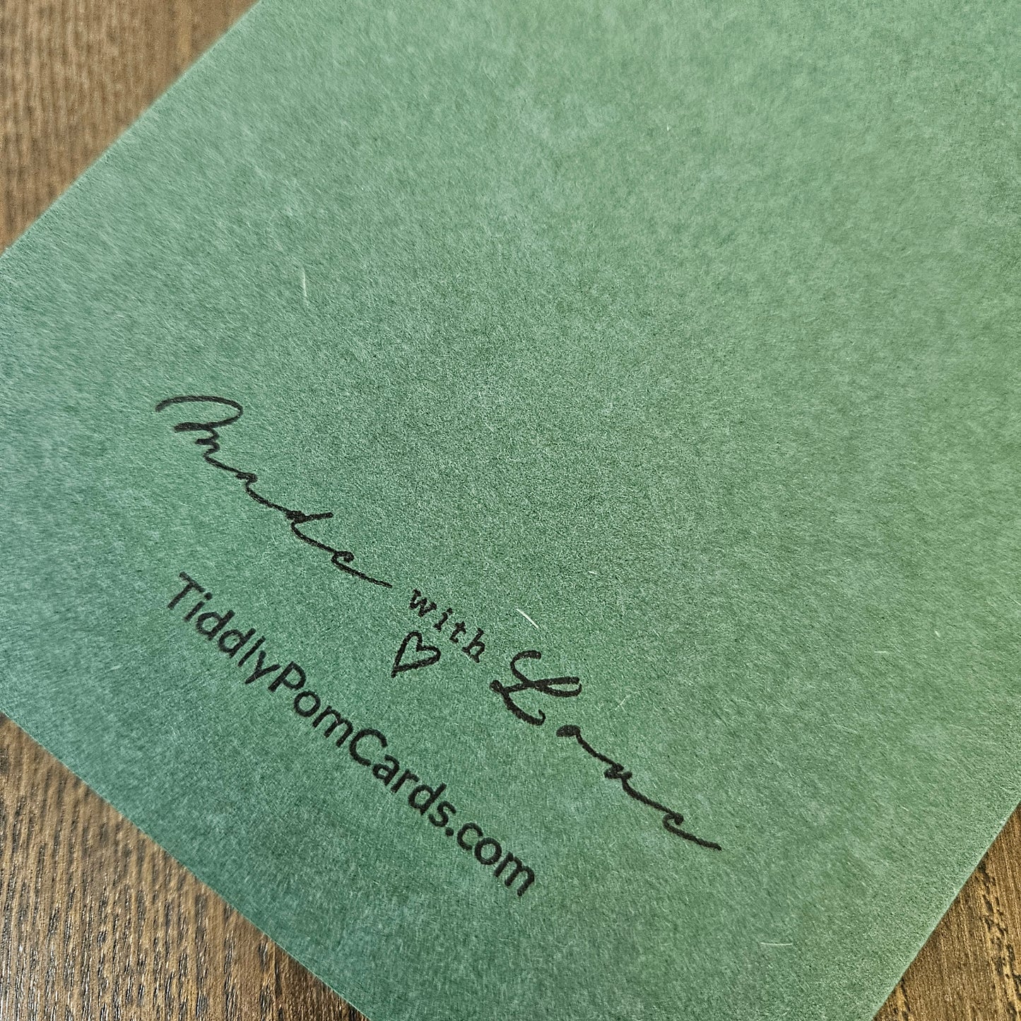 A close-up of a sage green textured card featuring the printed text 'Made with Love' and 'TidilyPomCards.com' in black ink, resting on a wooden surface.