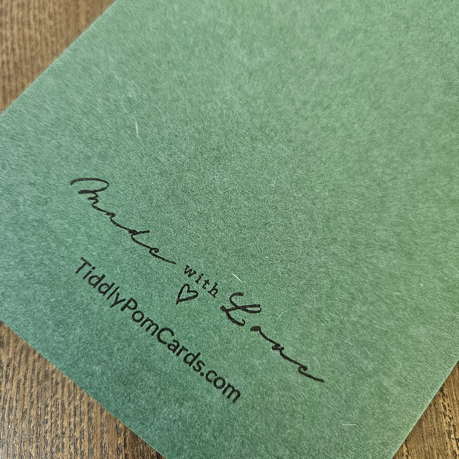 A close-up of a sage green textured card featuring the printed text 'Made with Love' and 'TidilyPomCards.com' in black ink, resting on a wooden surface.