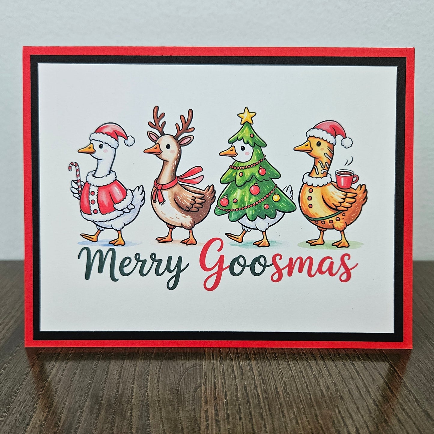 A square handmade greeting card with a red border, featuring four festive cartoon goslings dressed in Christmas attire on a white background, with the text 'Merry Goosmas' below. The card is displayed standing upright on a dark wooden table against a light gray wall.