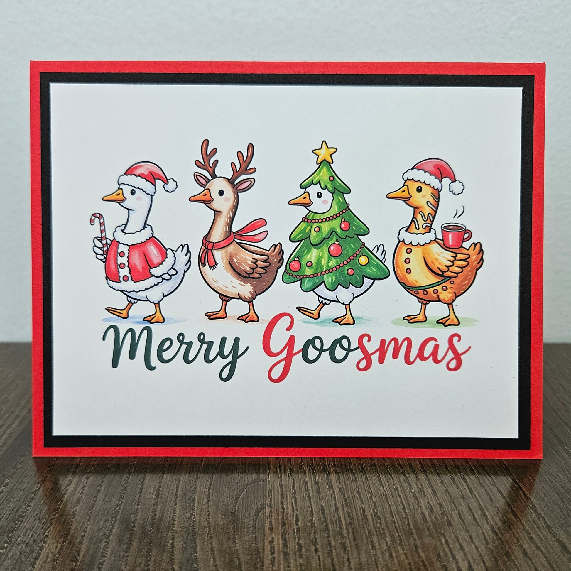 A square handmade greeting card with a red border, featuring four festive cartoon goslings dressed in Christmas attire on a white background, with the text 'Merry Goosmas' below. The card is displayed standing upright on a dark wooden table against a light gray wall.
